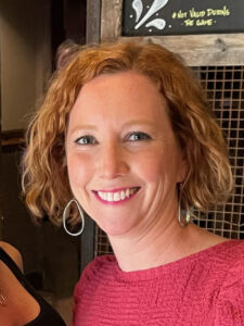 Copy Editor Monica Hodes-Smail smiles at the camera. She has short, reddish-blonde hair and wears silver hoop earrings and a red blouse.