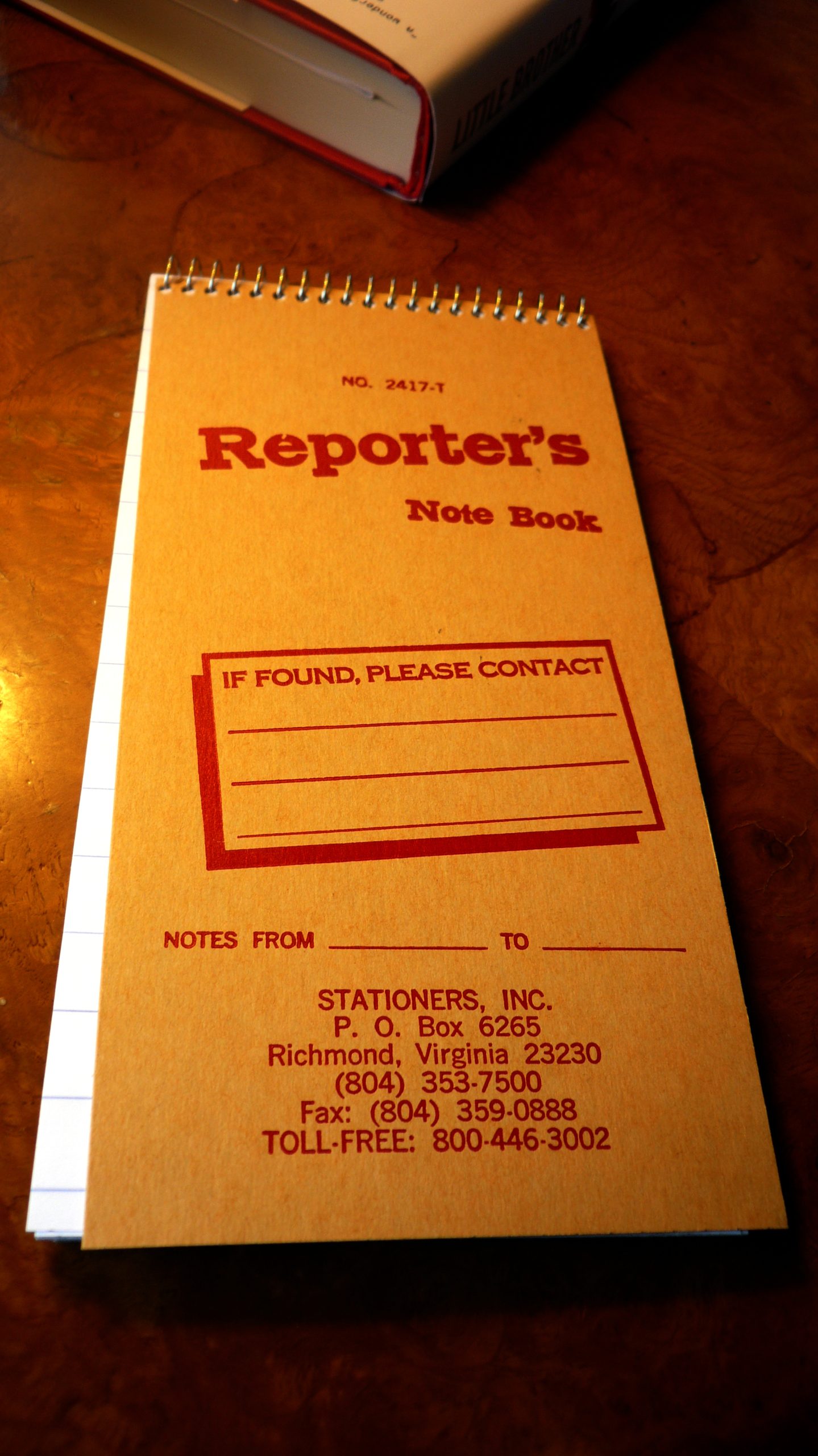 A long, thin notebook sits on a wood table near the corner of a book. The notebook cover is brown with red lettering that reads Reporter's Notebook.